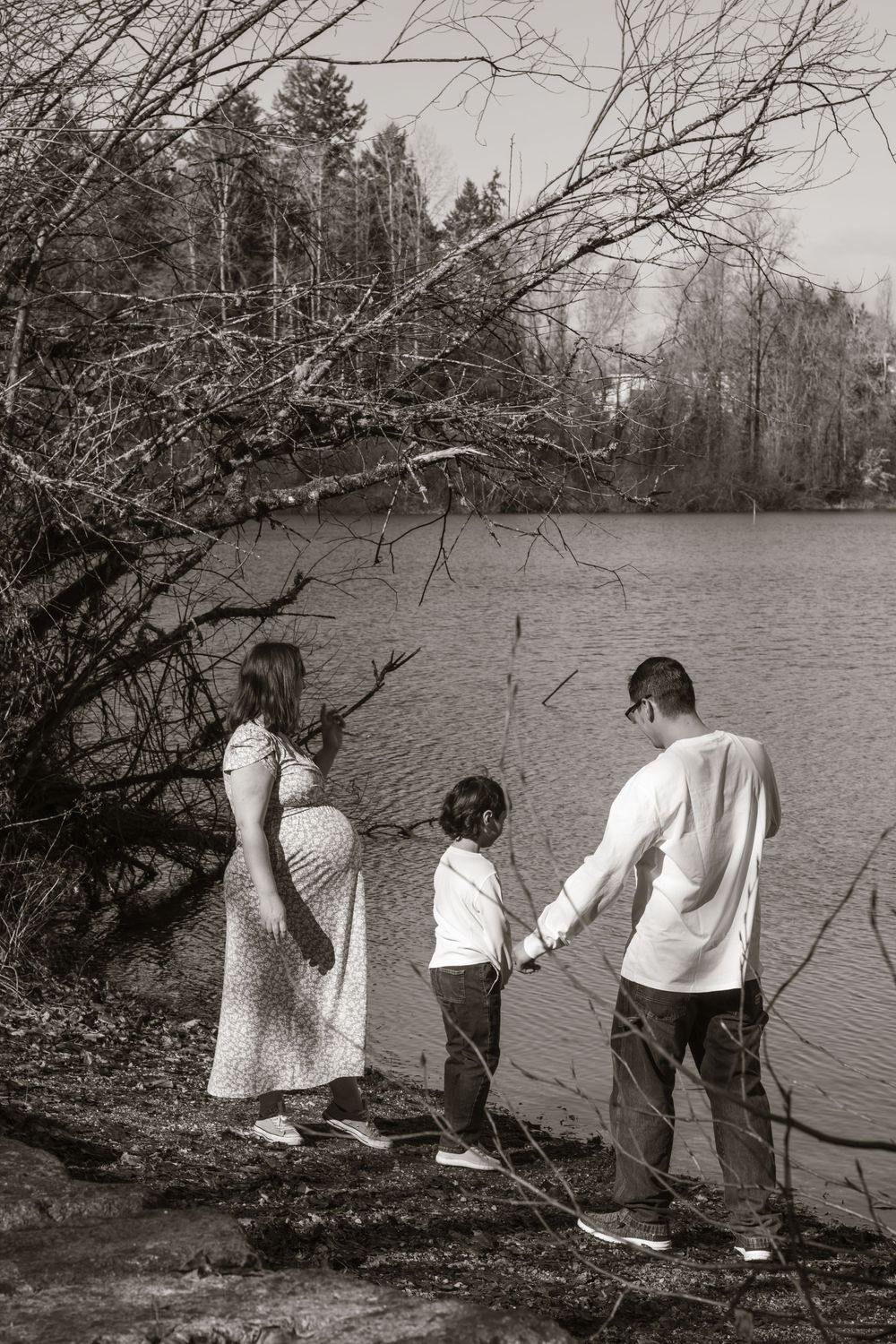 Black and white photo of a pregnant woman, child, and partner holding hands by a lakeside.