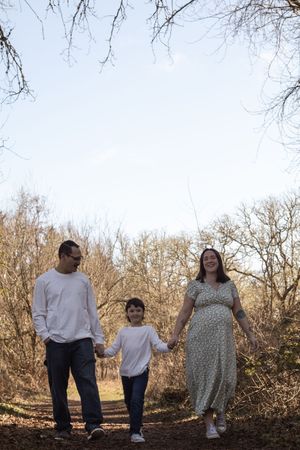 A pregnant woman in a floral dress walks hand-in-hand with her partner and child through bare trees.