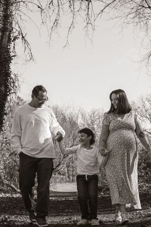 Black and white image of a laughing family with a pregnant mother walking together under bare trees.