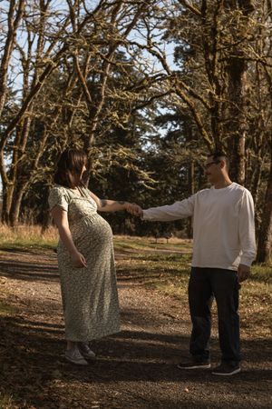 A pregnant woman in a floral dress holds hands with her partner on a sunlit forest path.