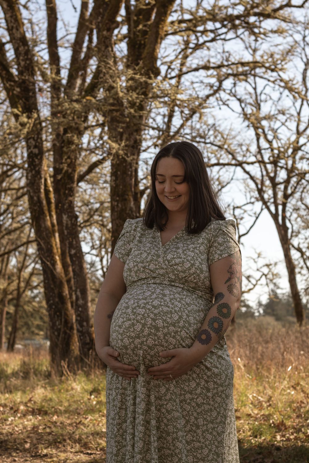 Smiling pregnant woman in floral dress cradling baby bump outdoors among bare trees in natural light.