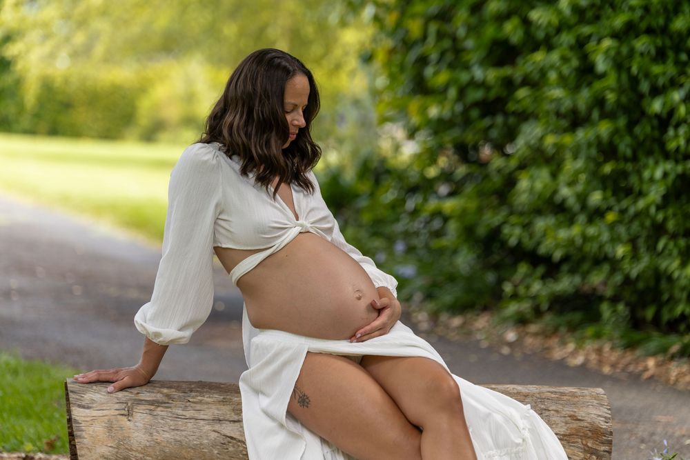 Expectant mother in white dress sits on rustic wooden bench during outdoor maternity photoshoot in nature setting.