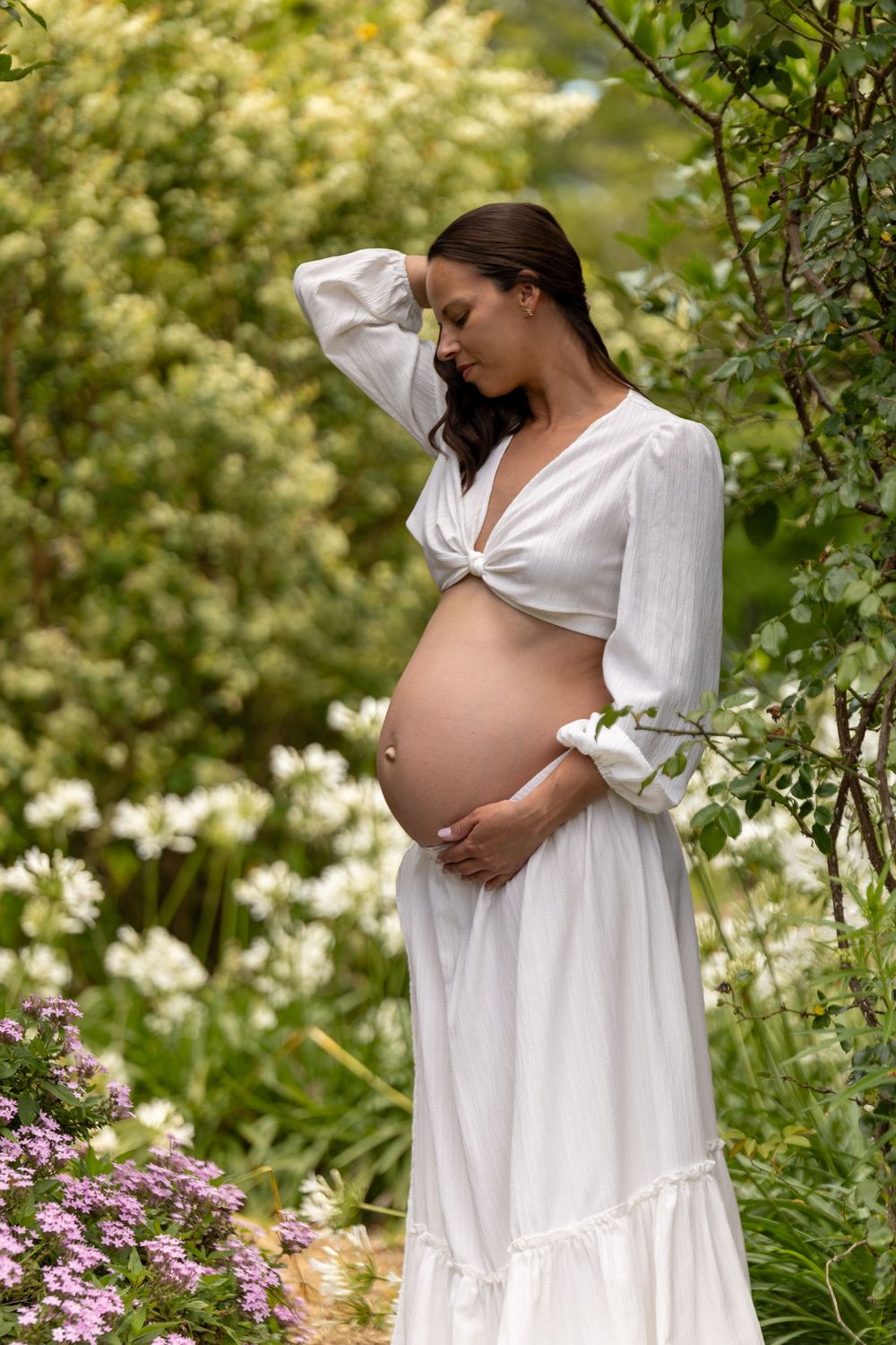 A serene maternity photoshoot in a garden with white flowing dress and pink flowers in the background.