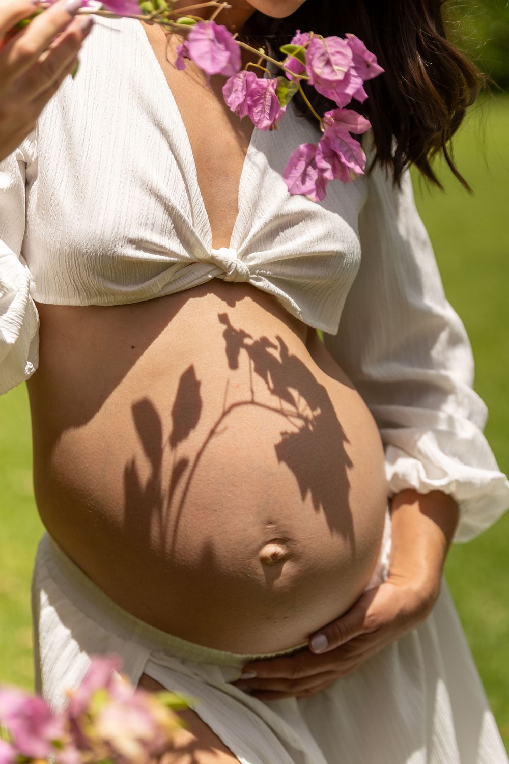Sunlit maternity photos with pink flowers creating delicate shadows on white fabric.