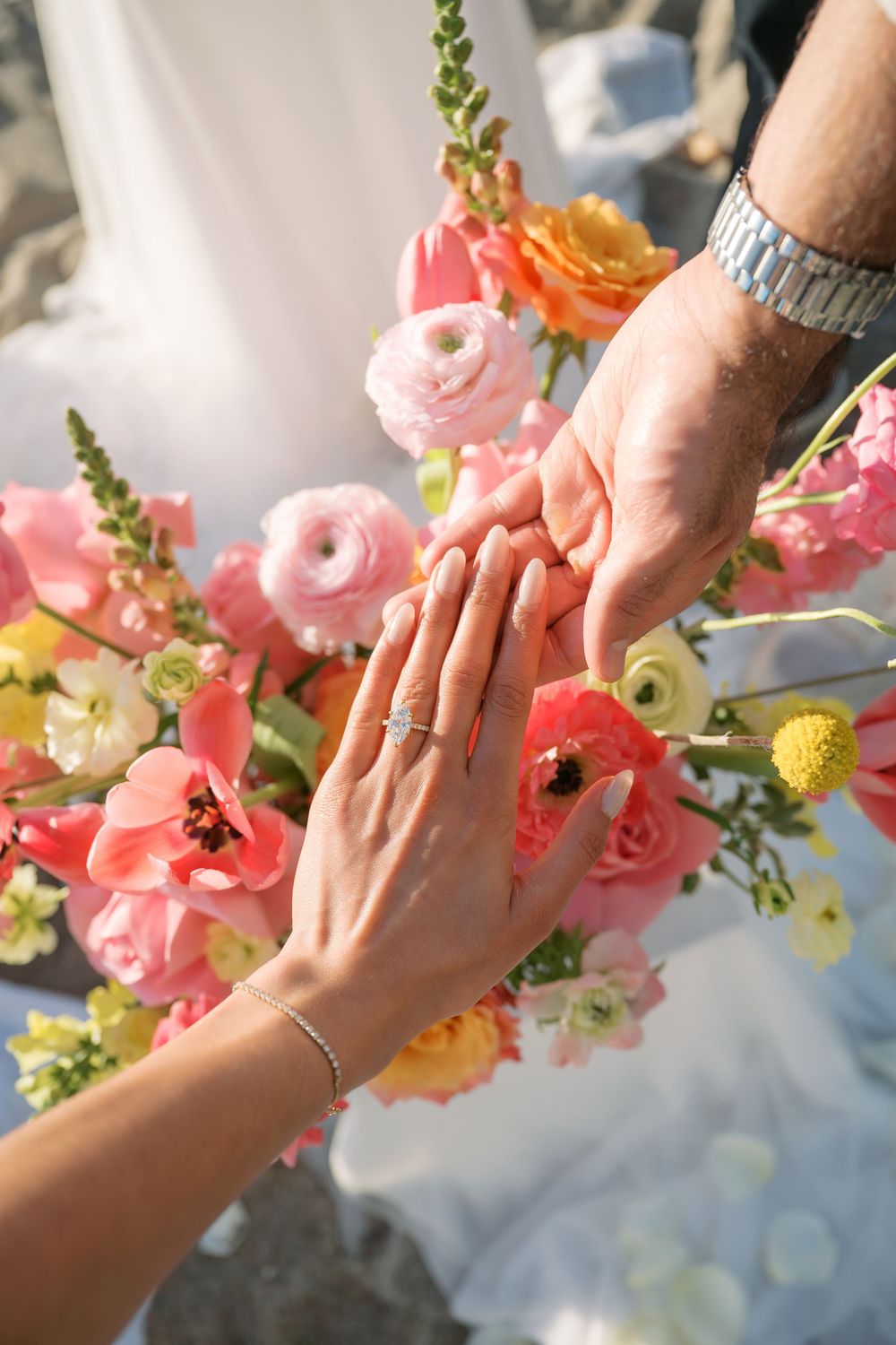 Two hands touching with engagement ring against beautiful pink and coral floral bouquet in sunlit setting.