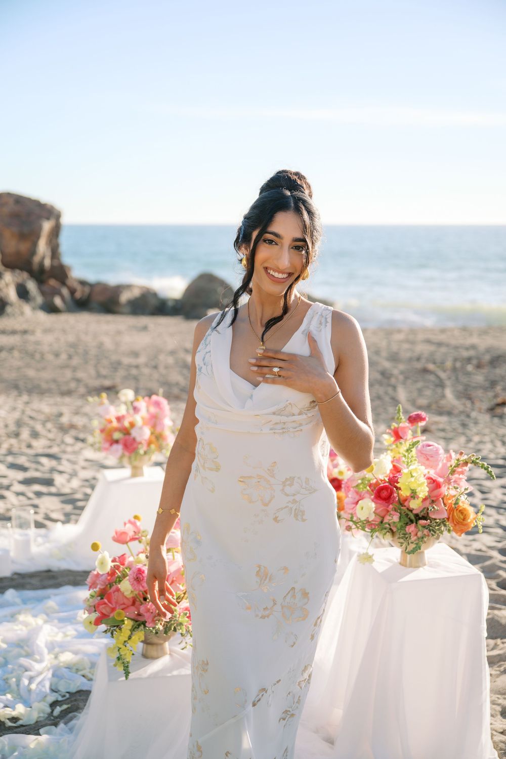 A romantic coastal bridal photoshoot featuring a white lace gown and vibrant coral and pink floral bouquet on a beach.