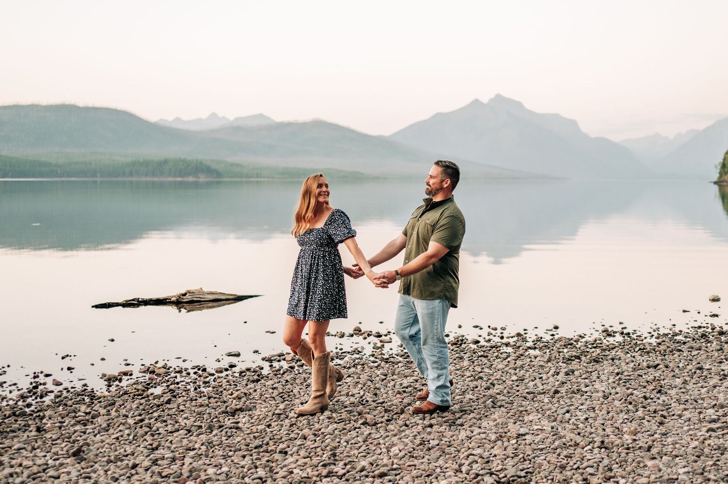 A couple walks along a scenic rocky beach with mountains and calm water in the background at sunset.