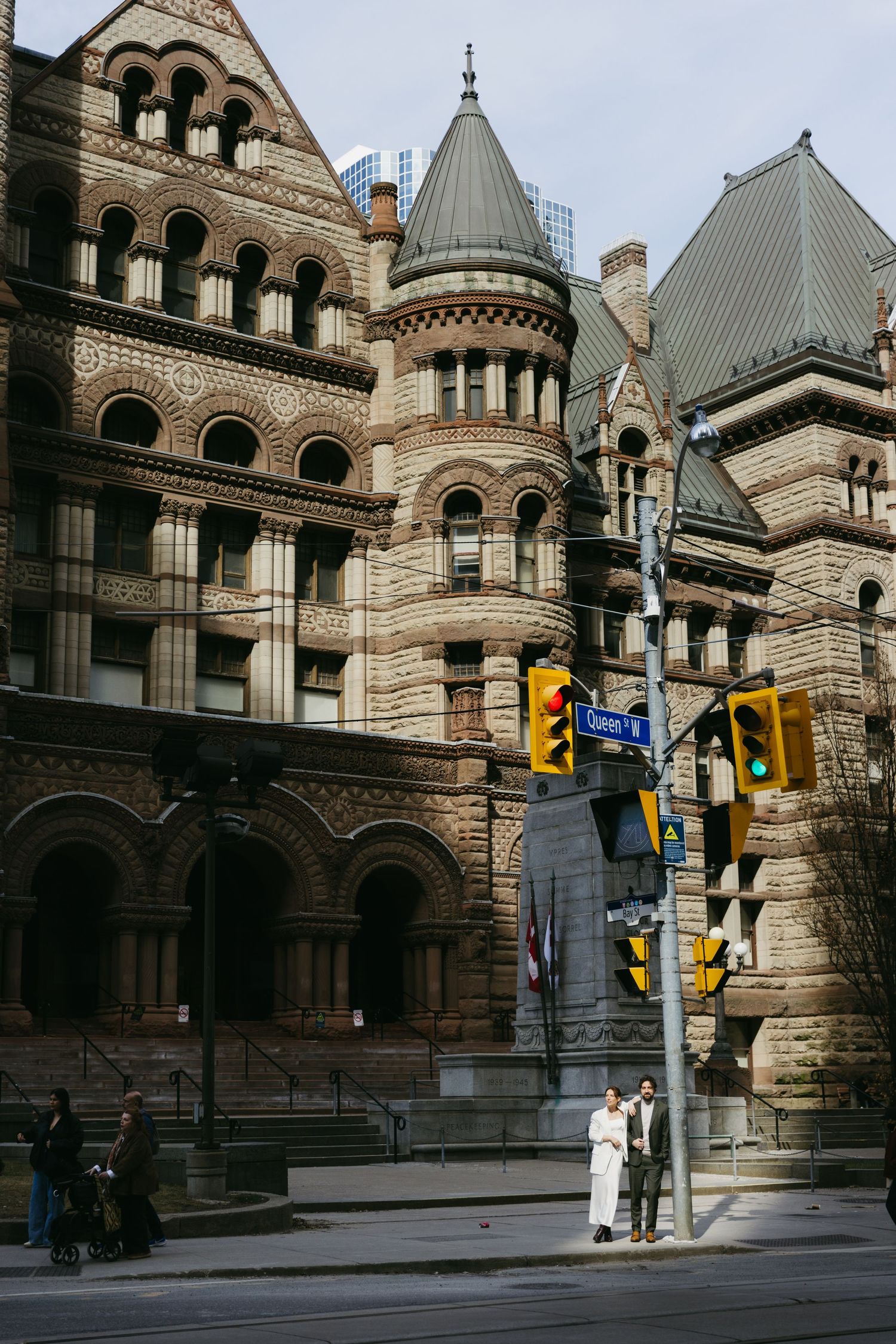 Wedding couple standing at Queen Street intersection beside historic stone building in downtown Toronto city setting.