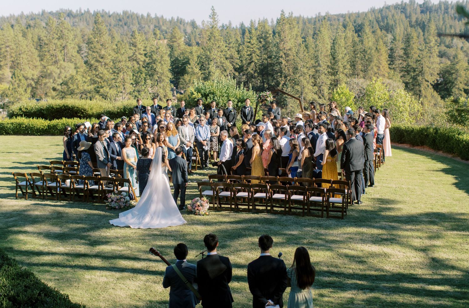 Outdoor wedding ceremony with guests seated in wooden chairs against forest backdrop.