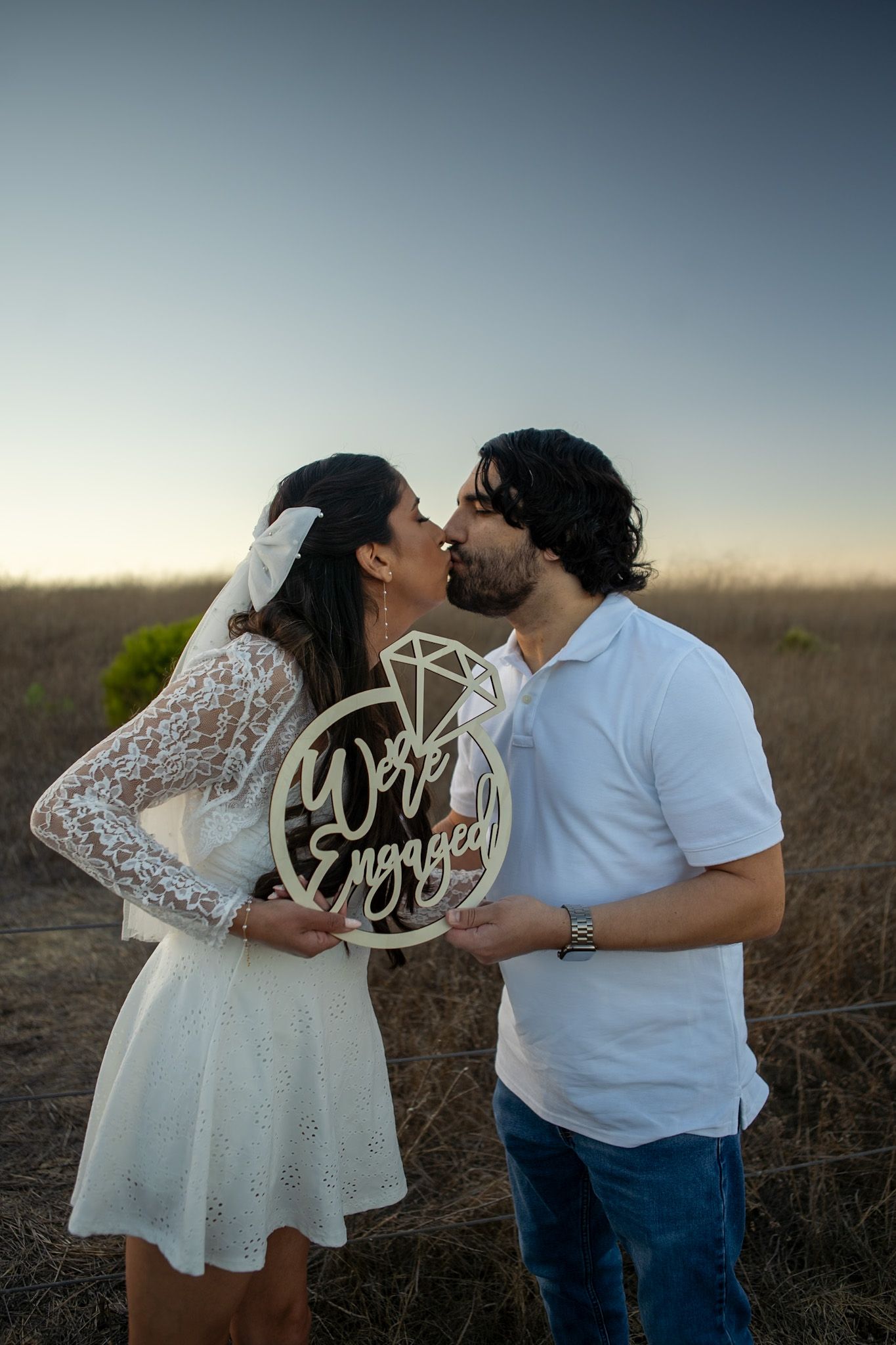 A couple shares a romantic moment at sunset while holding 'Mr' and 'Mrs' signs in a rustic outdoor setting.