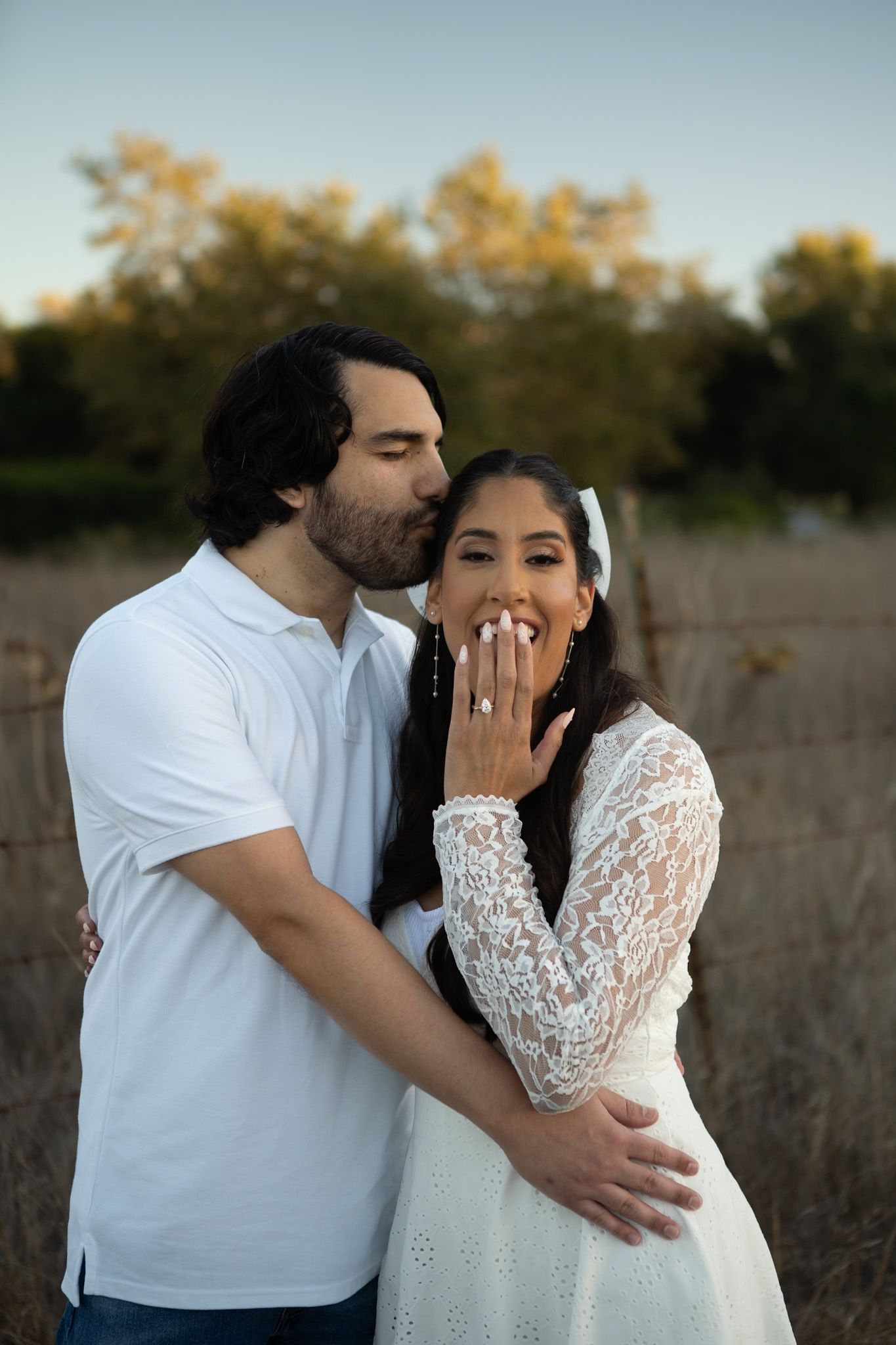 A couple shares an intimate moment in a field at sunset, with the woman wearing a white lace dress.