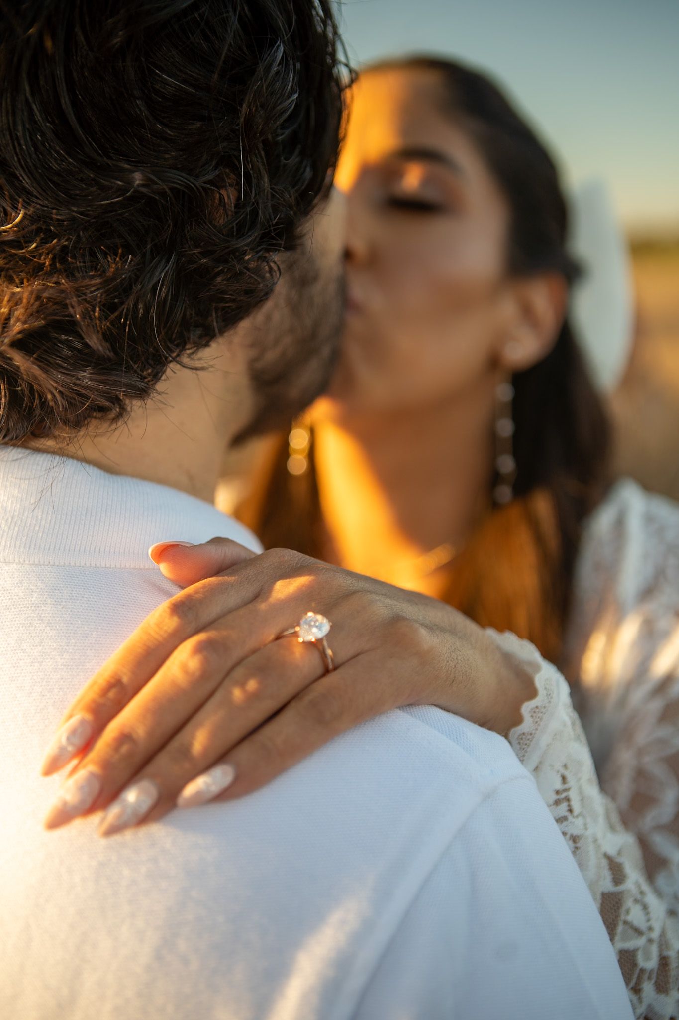 A couple shares an intimate moment at sunset with a diamond ring visible on her hand.