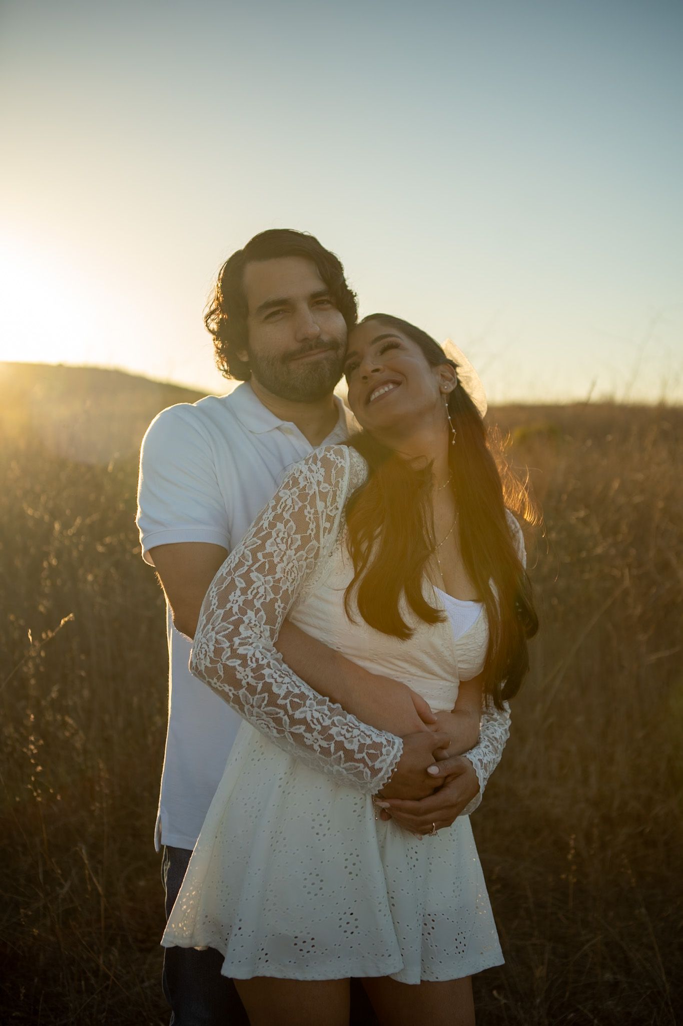 A couple in white outfits share intimate moments during a golden hour photoshoot in a field at sunset.