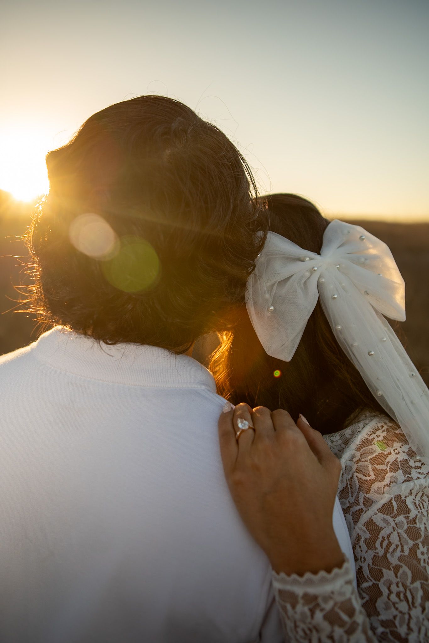 Romantic couple embraces during golden hour sunset, with bride wearing white lace and a bow in her hair.