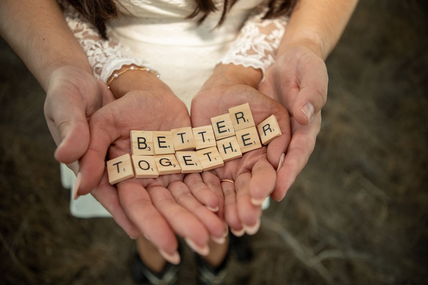 Hands holding wooden Scrabble tiles spelling out 'BETTER TOGETHER' against a white dress.