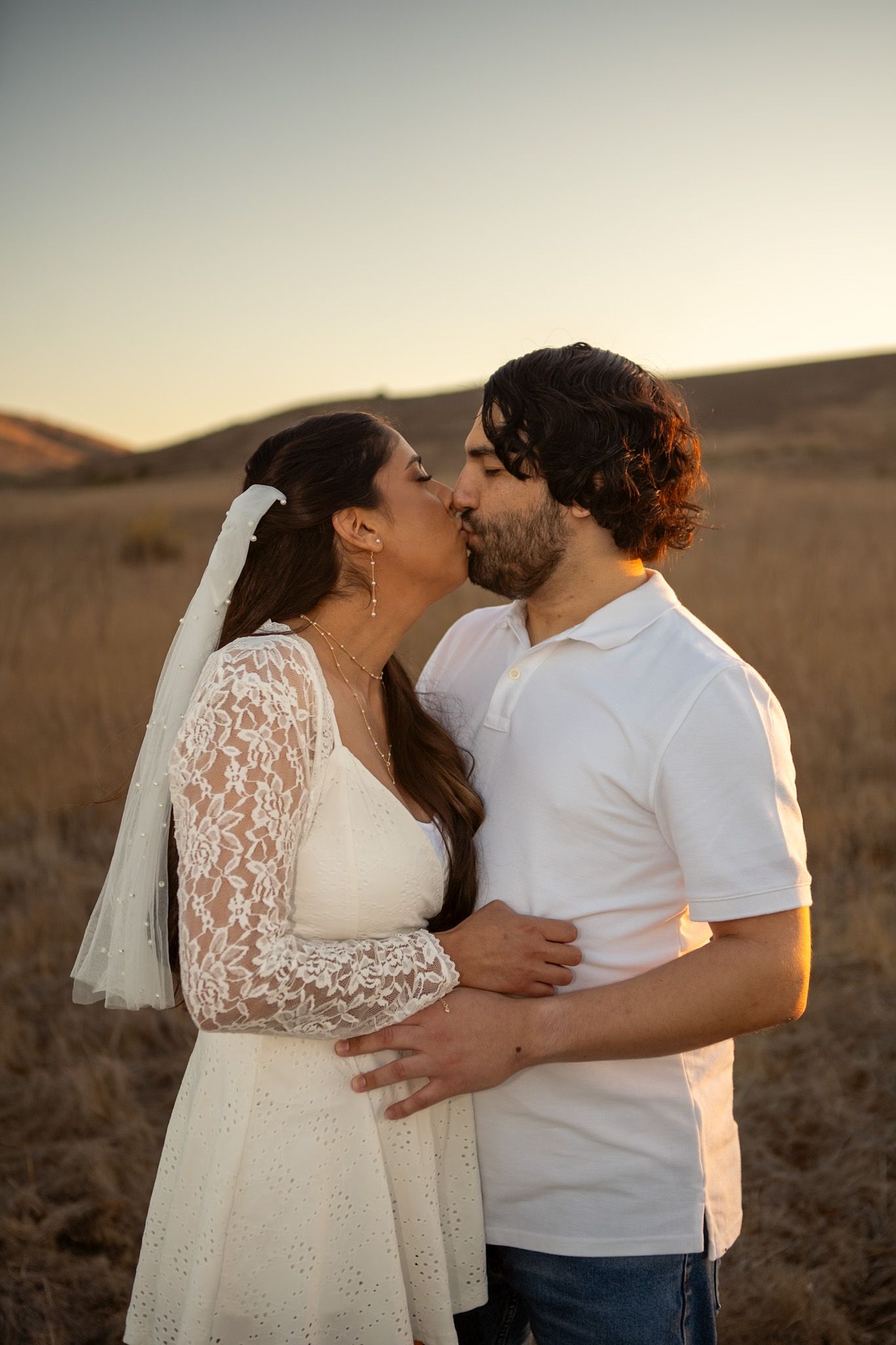 A couple shares an intimate moment at sunset in a desert landscape while wearing white wedding attire.