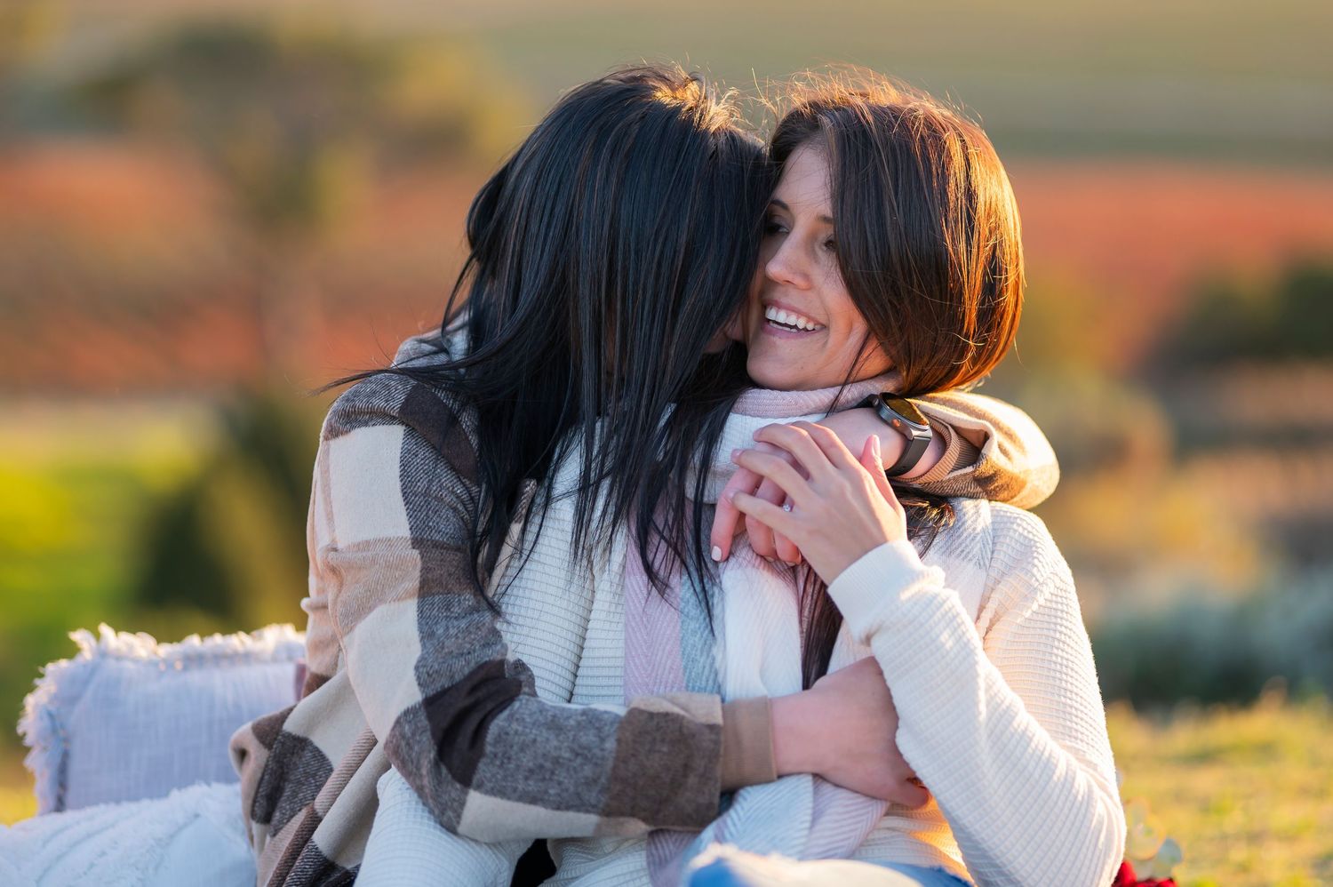 Two young women with dark hair share a warm embrace outdoors, smiling joyfully in golden sunlight.