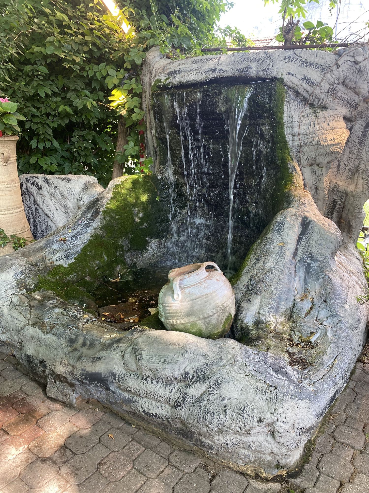 A stone garden water feature with cascading waterfall and decorative ceramic pot on brick paving.