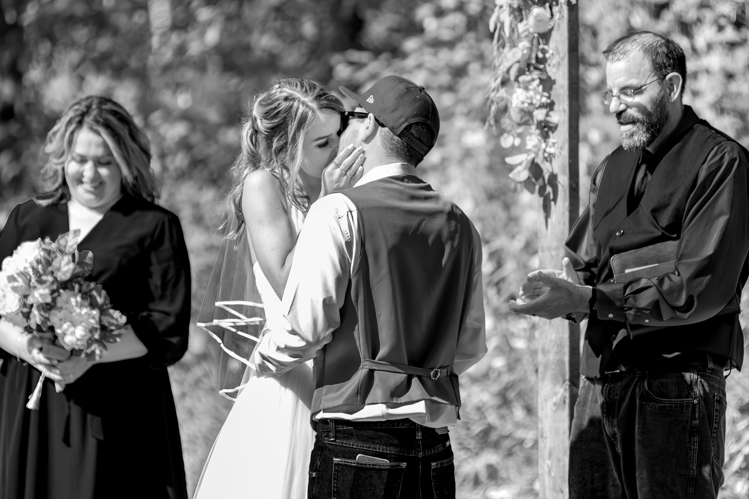 Black and white photo of an outdoor wedding ceremony with a couple and officiant under trees.