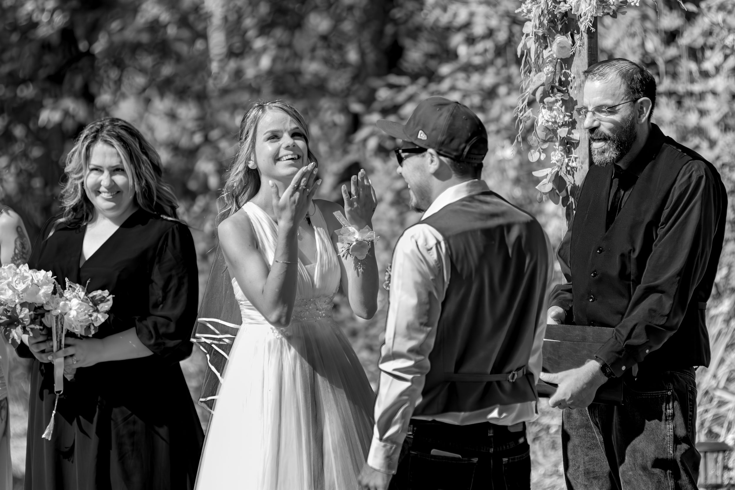 Black and white candid photo of a joyful outdoor wedding ceremony with people gathered under trees.