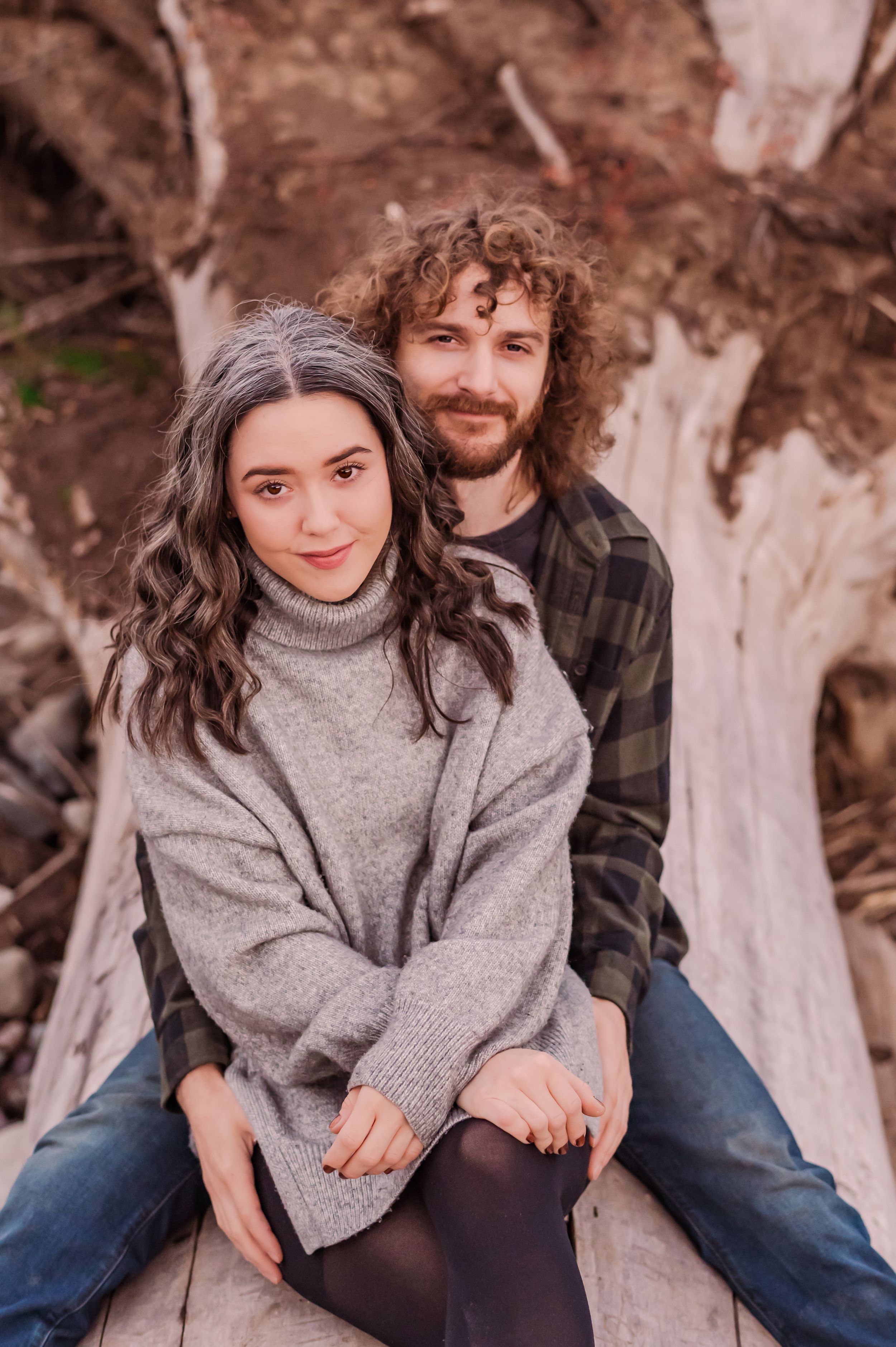 A couple sits together outdoors wearing cozy sweaters against a natural backdrop with tree trunks.