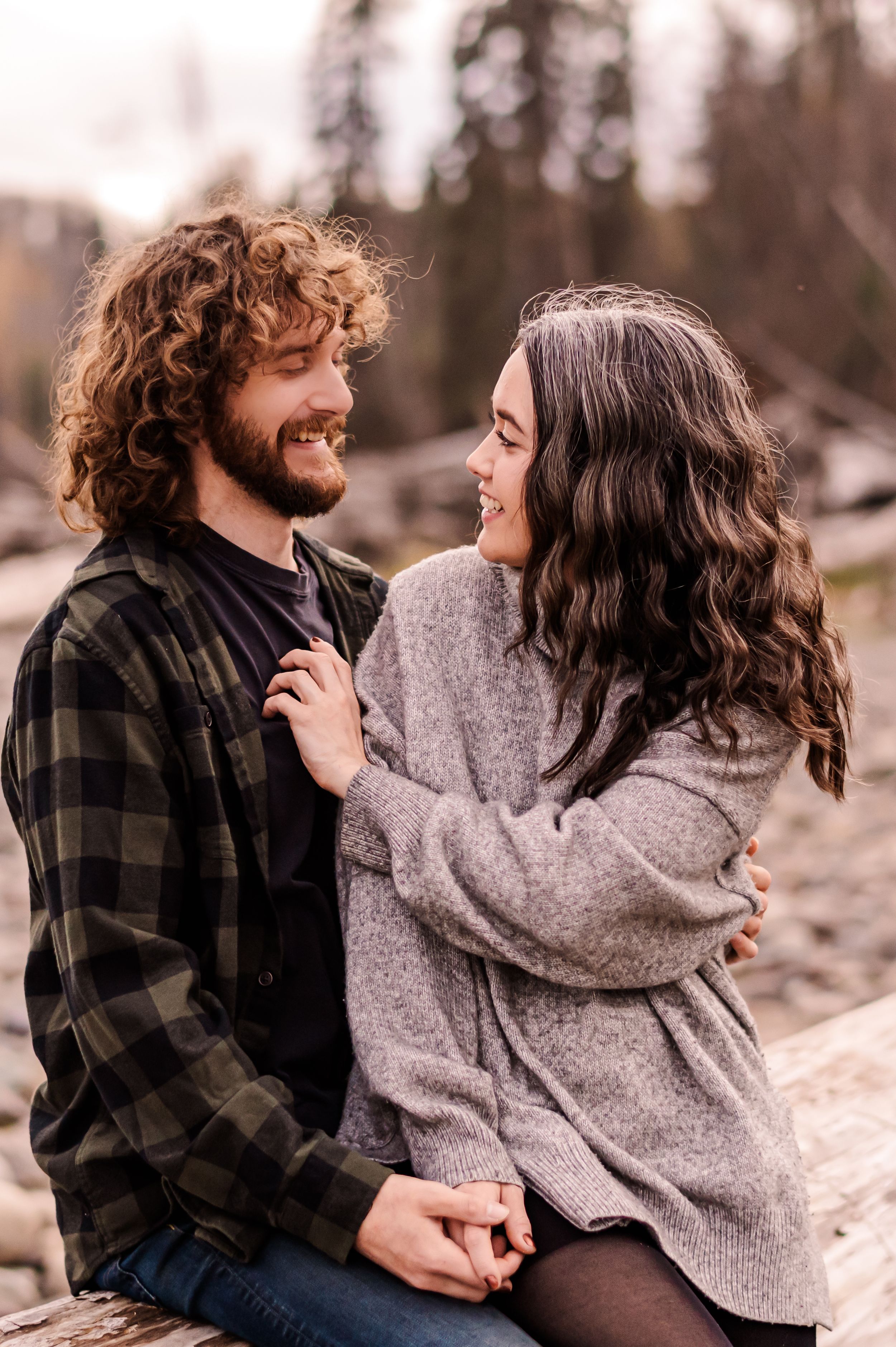 A couple laughs and embraces while sitting together outdoors, wearing cozy sweaters in a romantic autumn setting.