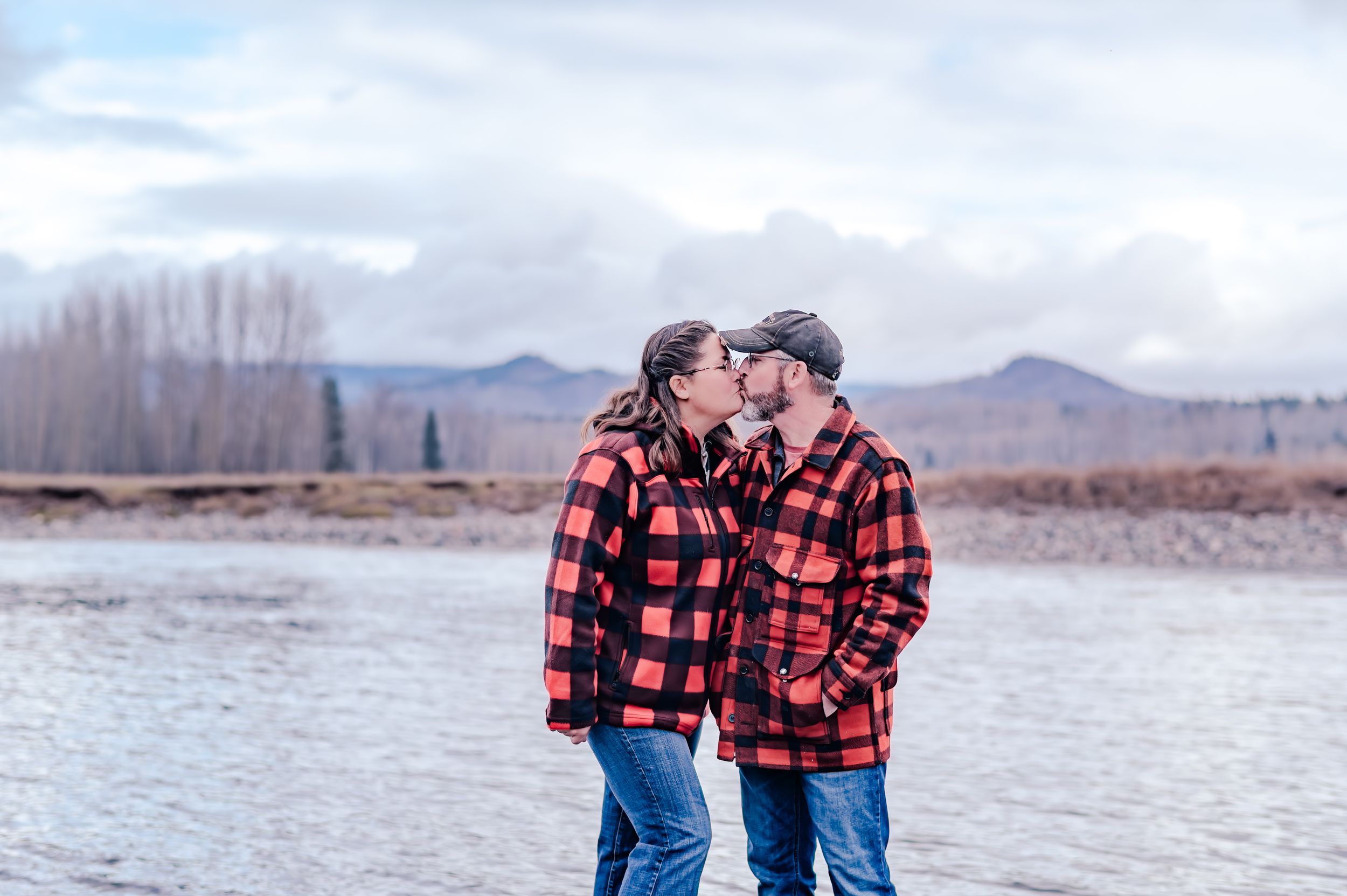 Two people in matching buffalo plaid jackets share a tender moment by a lake with mountains in the background.