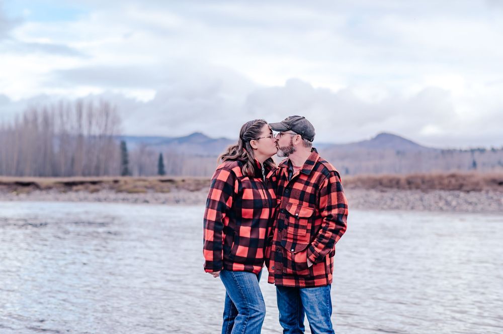 Two people in matching buffalo plaid jackets share a tender moment by a lake with mountains in the background.