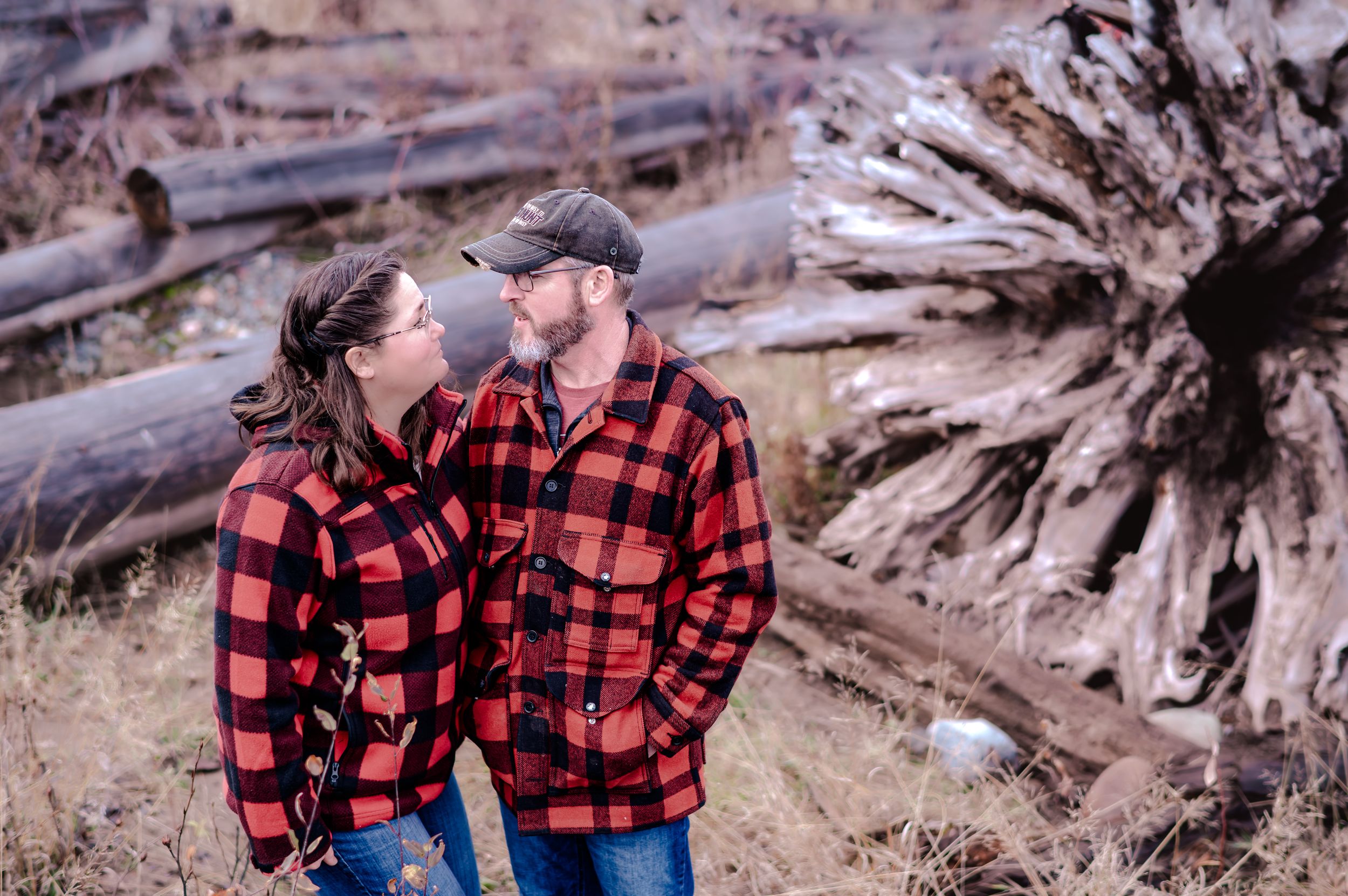 Couple in matching red buffalo plaid flannel shirts embrace near driftwood on a beach during sunset.