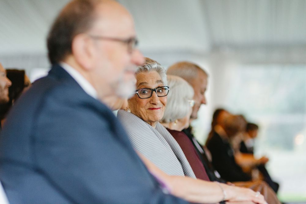 Elderly woman with glasses smiling at a formal outdoor event, seated among other attendees in elegant attire.