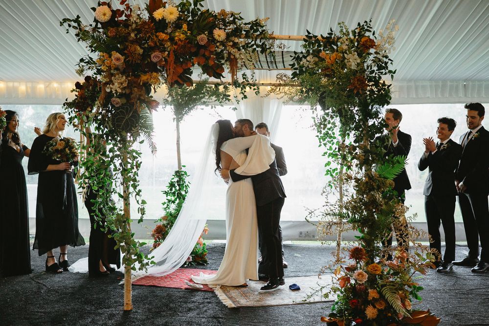 Wedding ceremony under white tent decorated with vibrant autumn floral arrangements and greenery.