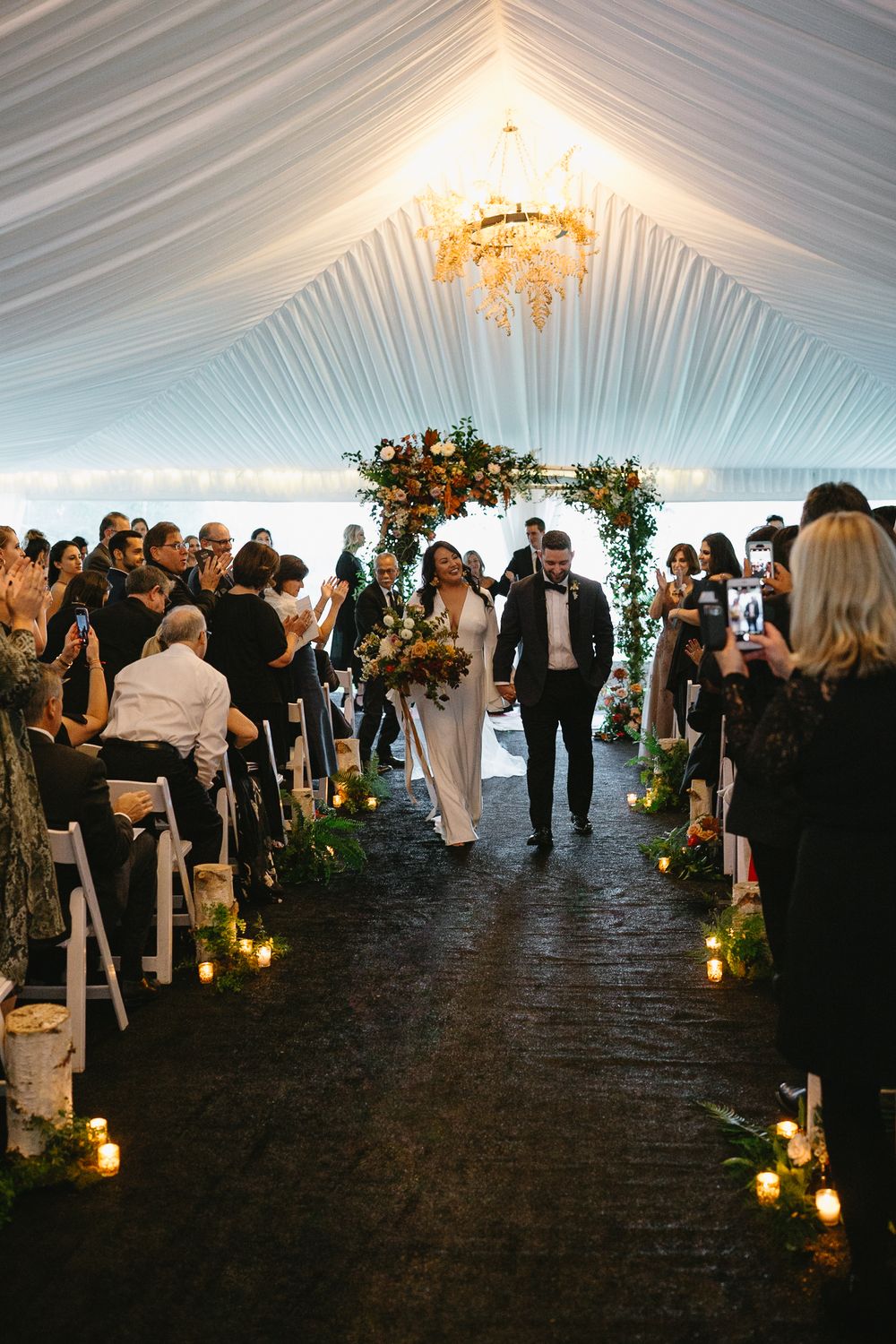 Guests gather inside an elegant white tent for an evening wedding ceremony illuminated by glowing candles along the aisle.