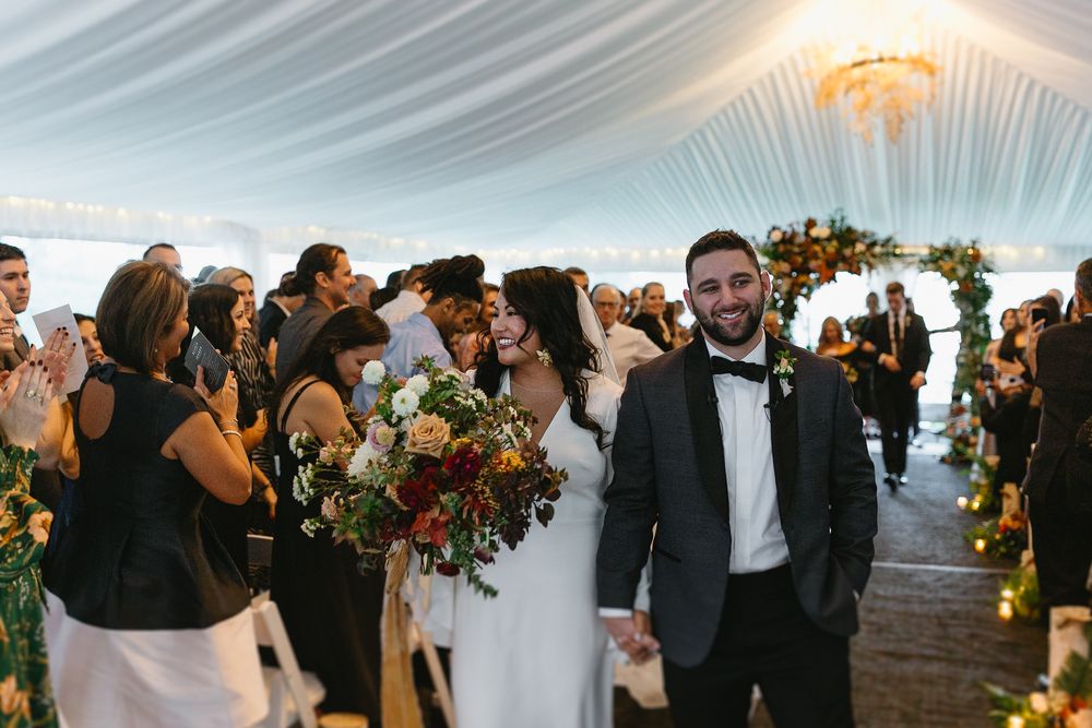 Wedding guests gather on a beach at sunset under a dramatic cloudy sky during a seaside celebration.