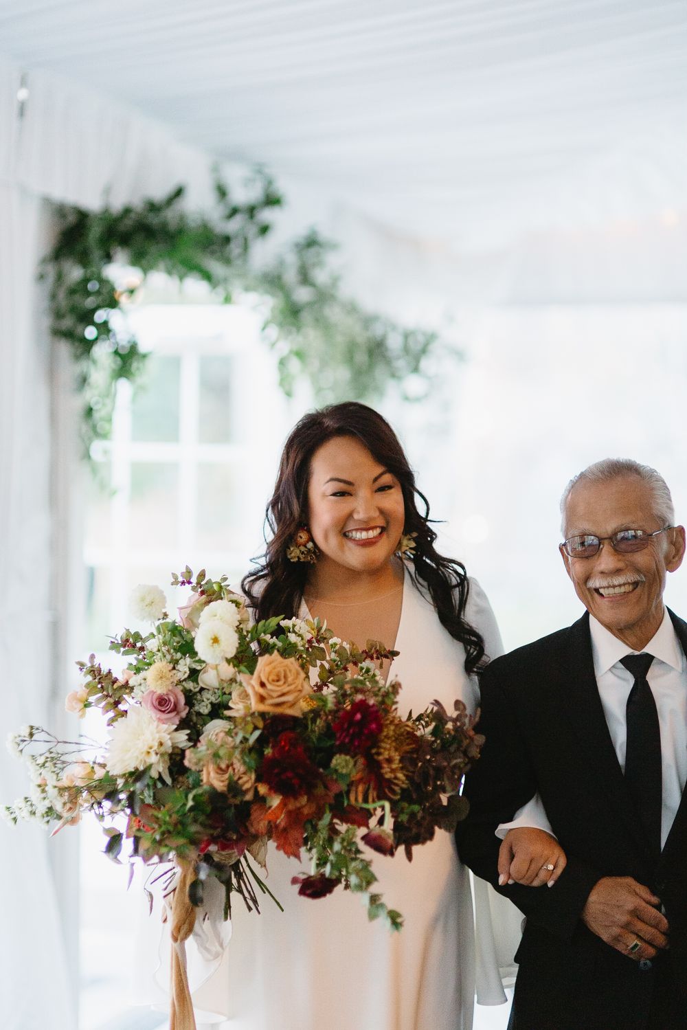 A joyful wedding party walks down a white-columned hallway carrying vibrant fall-colored floral bouquets.