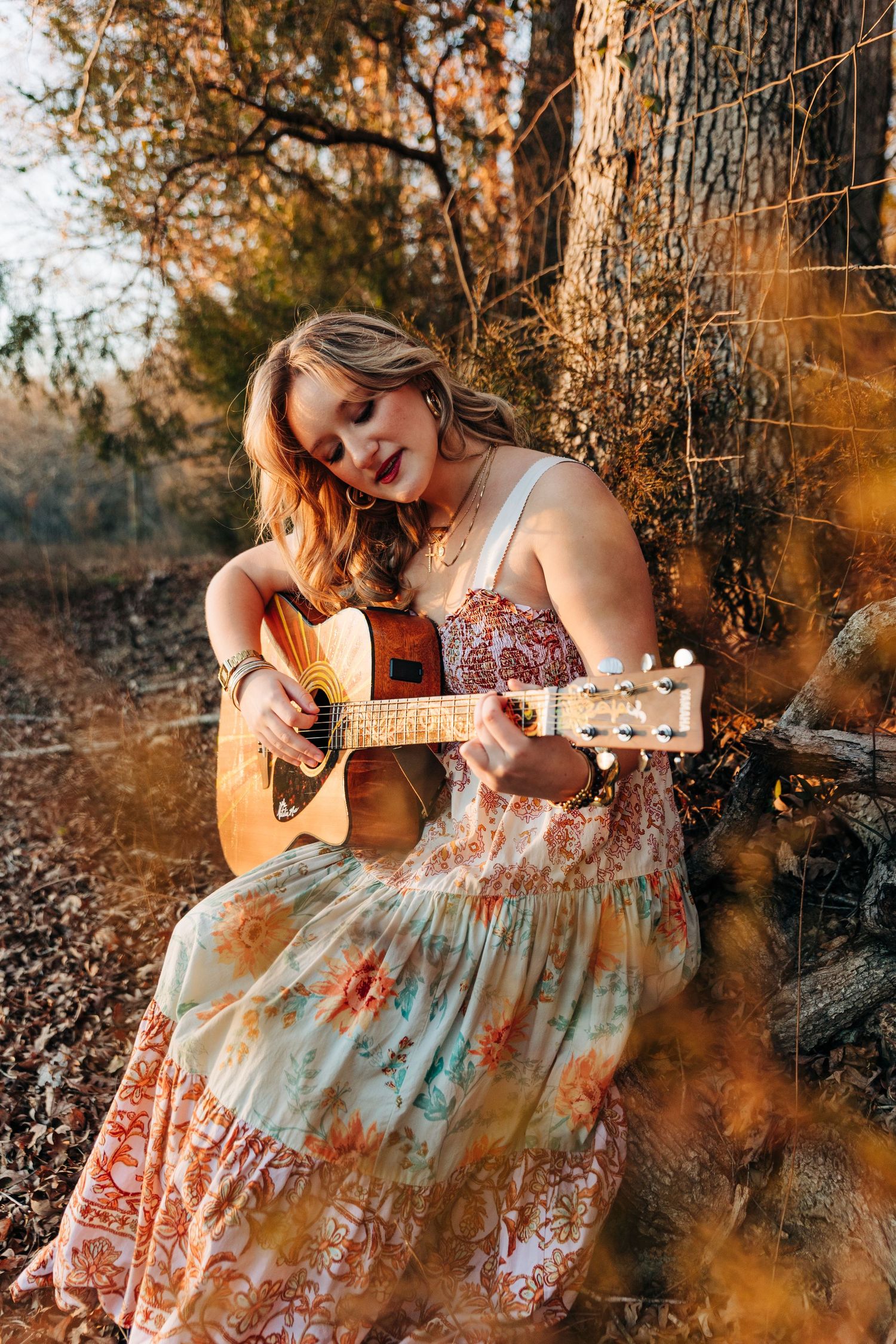 A musician in a floral dress plays acoustic guitar while sitting against a tree trunk in warm autumn lighting.