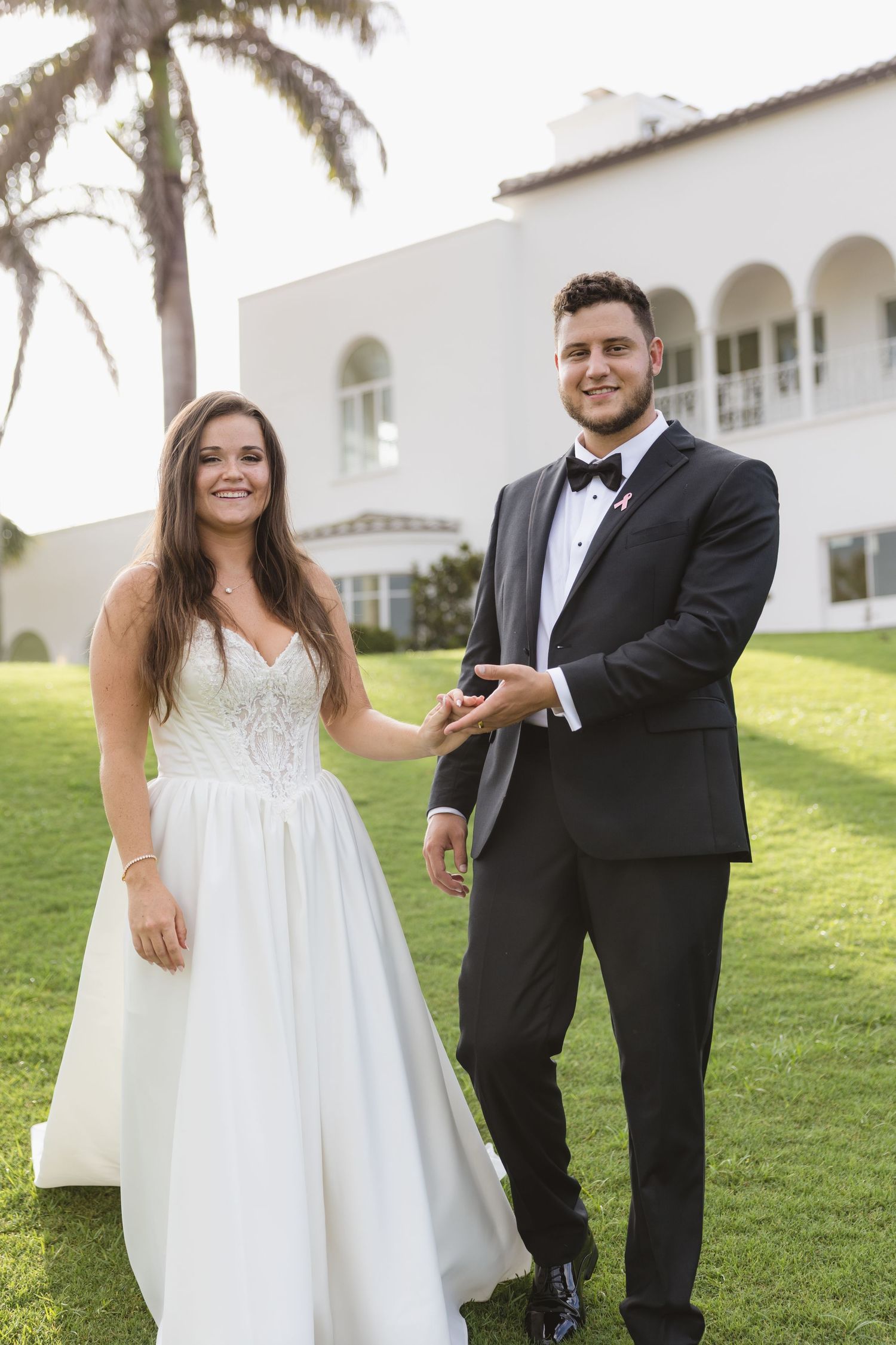 A couple in white and beige attire dances joyfully on grass while guests throw confetti during an outdoor celebration.