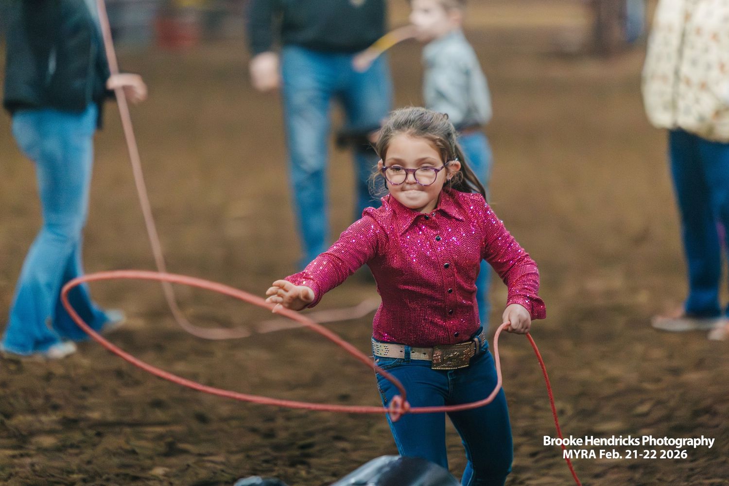 A child in a pink sweater and glasses plays with a lasso on muddy ground during an outdoor activity.