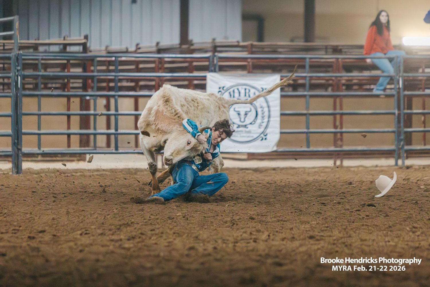 A bull rider in turquoise attire gets thrown off during a rodeo competition in an indoor arena.