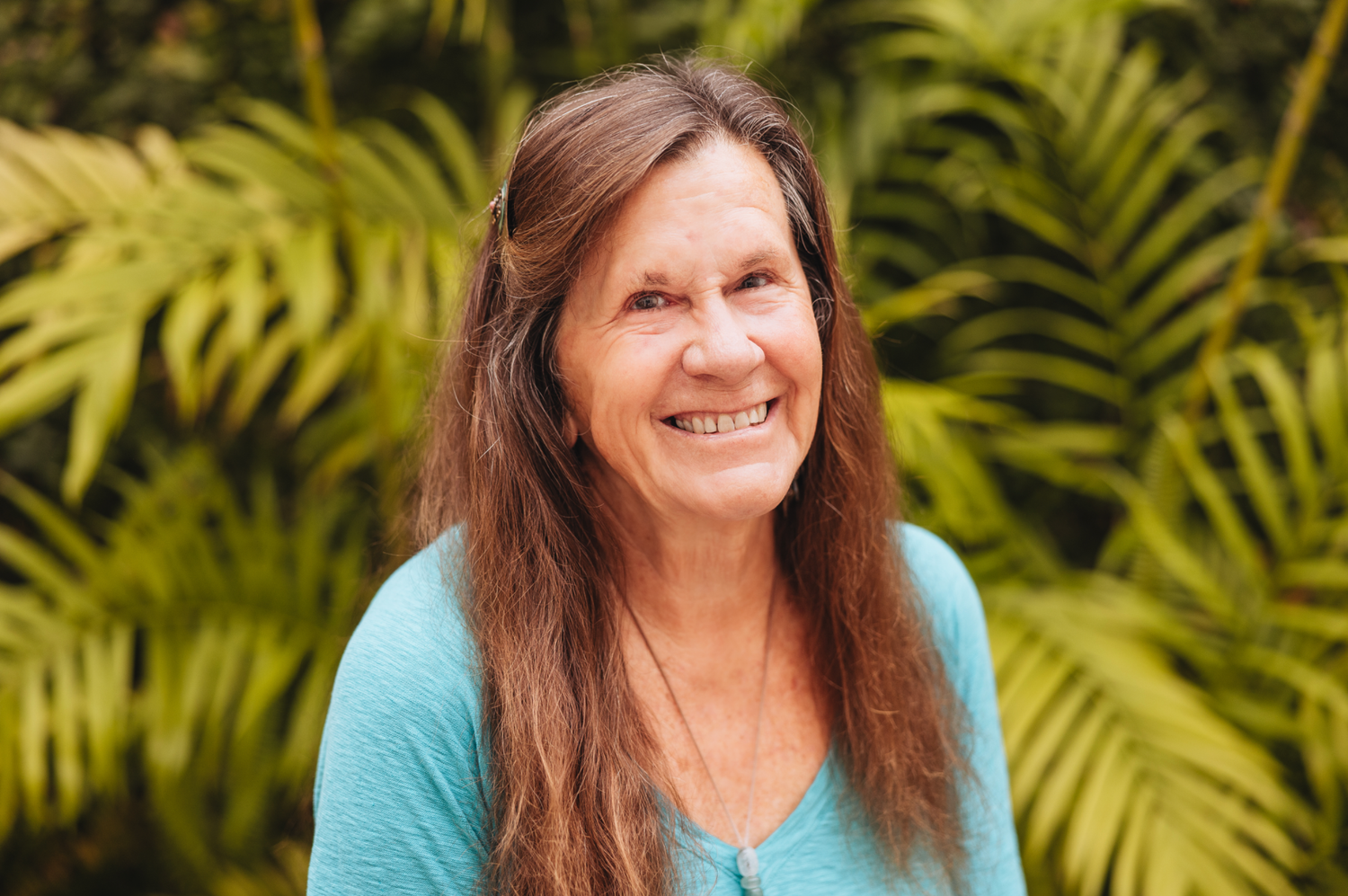 Woman with long brown hair wearing turquoise top smiles warmly against tropical foliage background.
