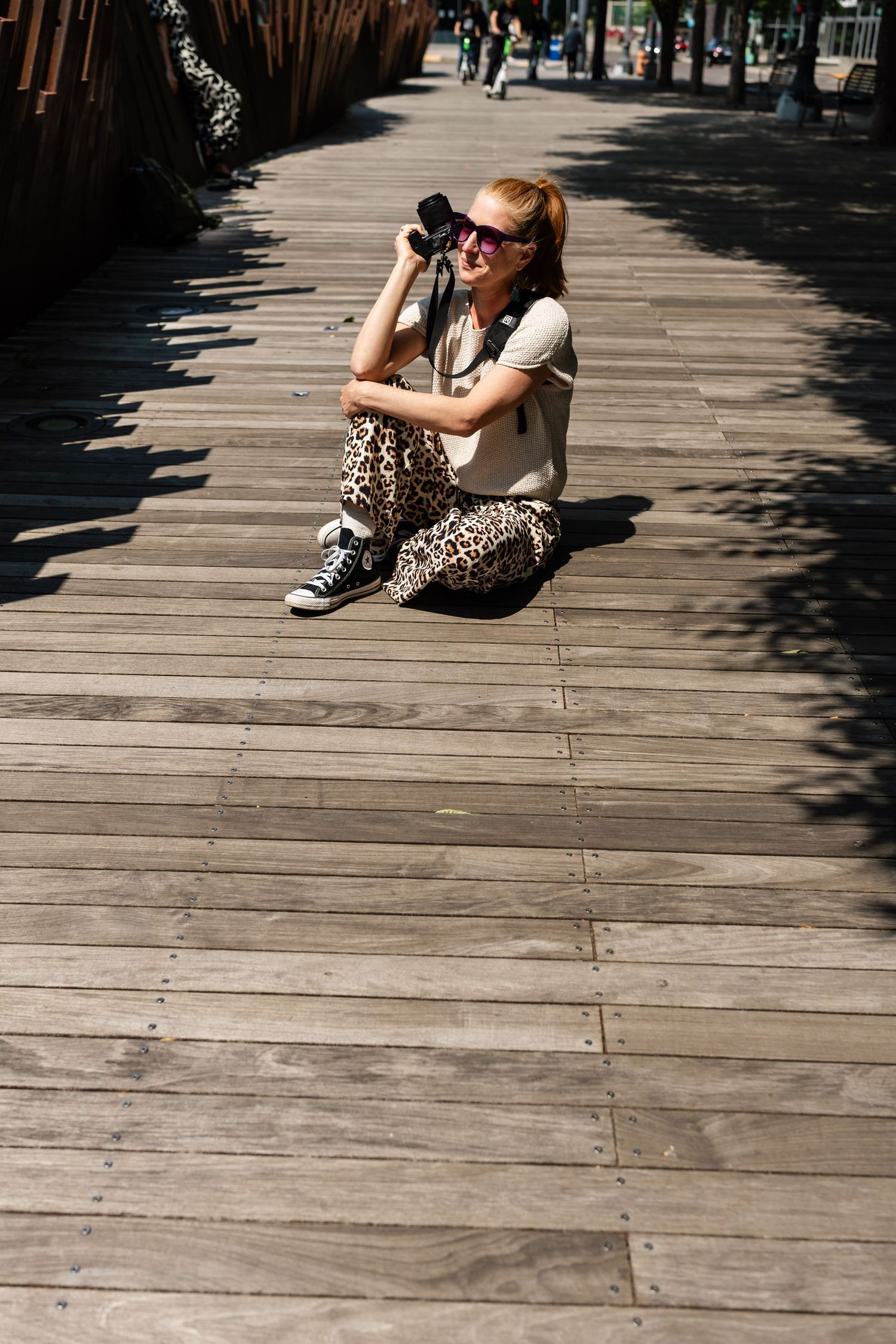 A photographer crouches low on wooden boardwalk planks to capture a shot in bright sunlight.