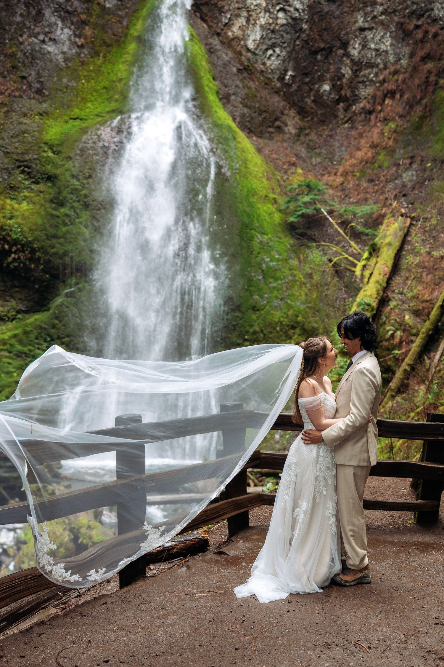 A couple in wedding attire poses romantically with a flowing veil in front of a majestic waterfall in a forest setting.