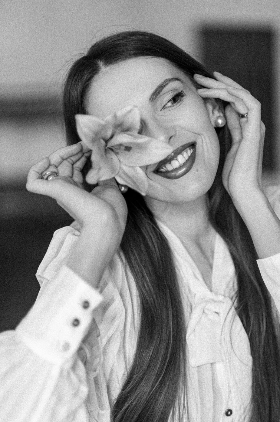 Black and white portrait of a joyful person with long dark hair wearing a white button-up shirt, laughing with hands near face.