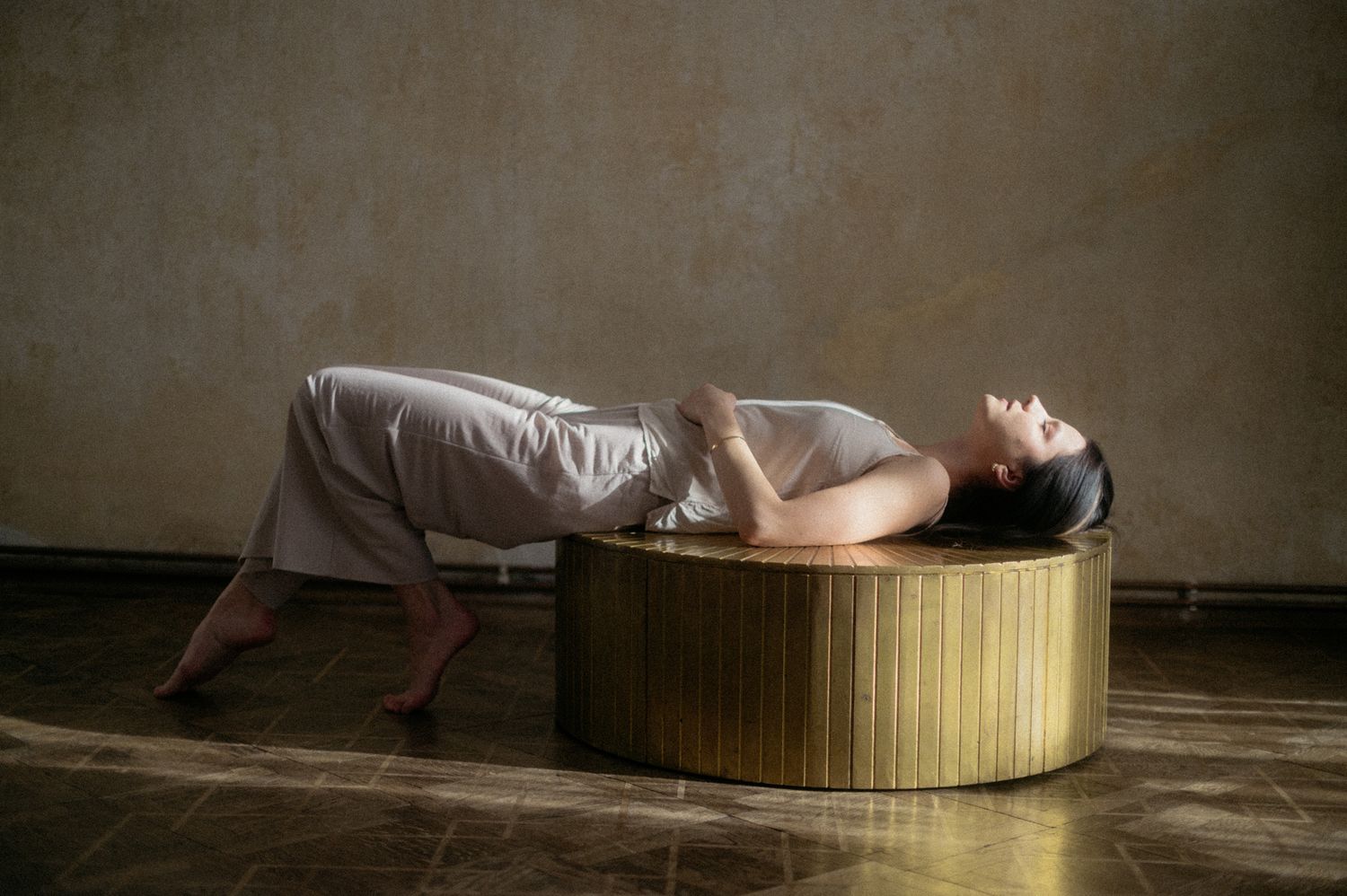 A person in white clothing lies arched backwards across a cylindrical bamboo platform in a minimalist setting.