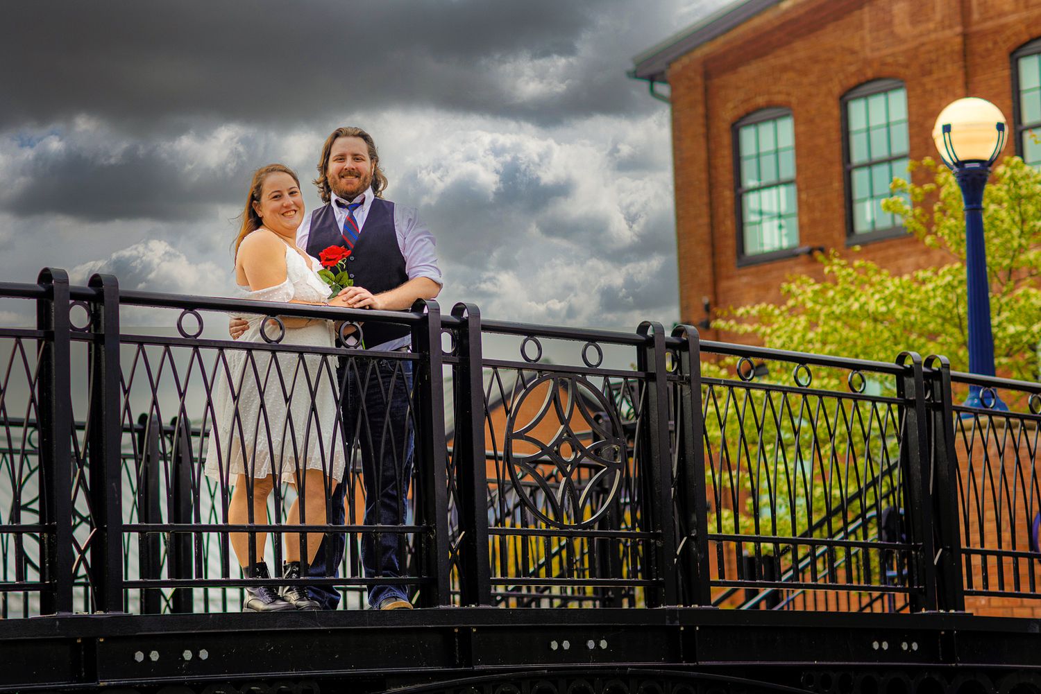 A decorative black metal fence with ornate details stands against a dramatic cloudy sky near brick buildings.