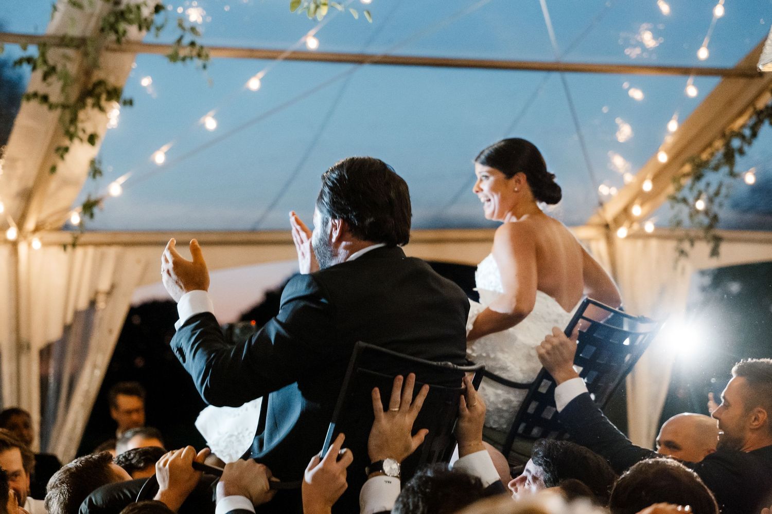 Wedding guests dance and celebrate under twinkling string lights at an evening reception.