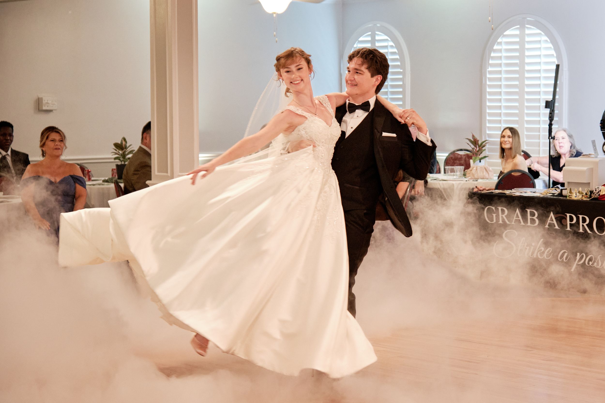 Bride and groom share first dance on foggy dance floor, bride in white gown, groom in black tuxedo at wedding reception.