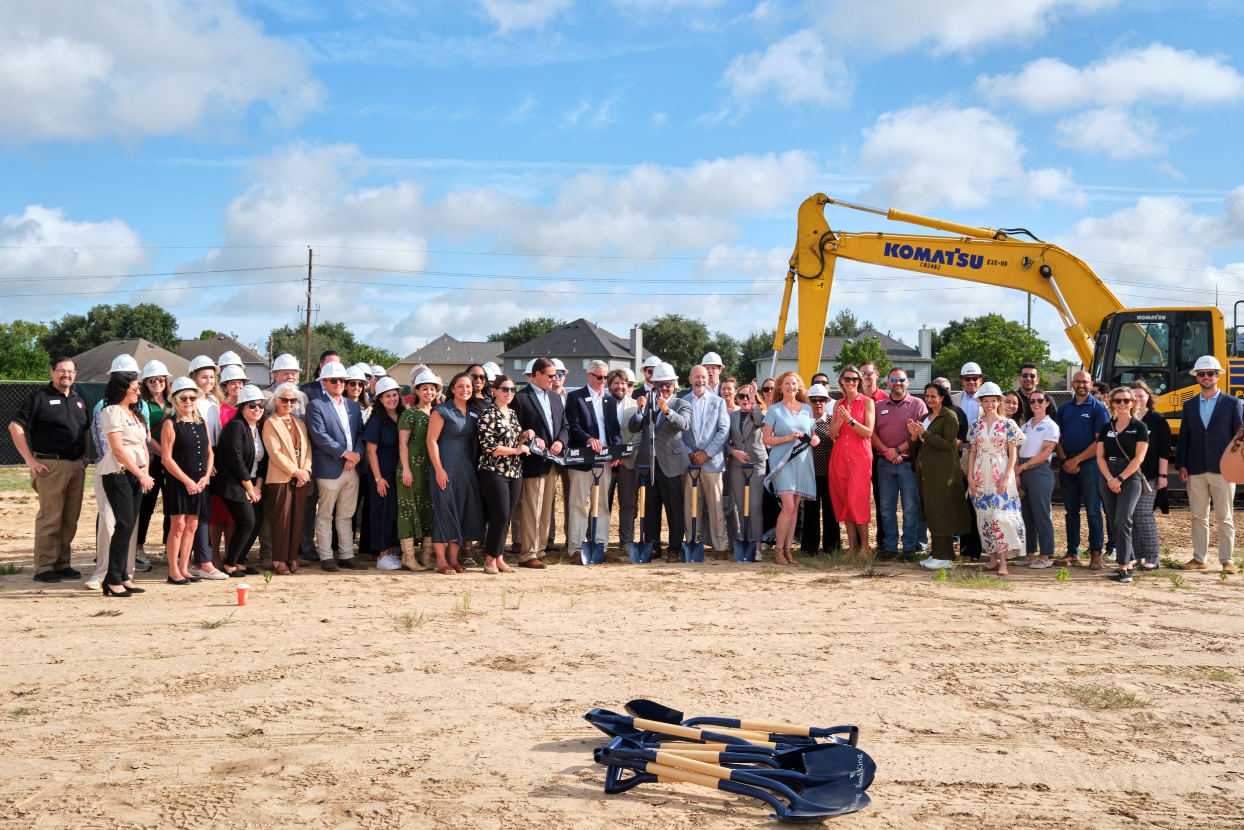 A large group of people in business attire gather for a groundbreaking ceremony with a yellow excavator behind them.