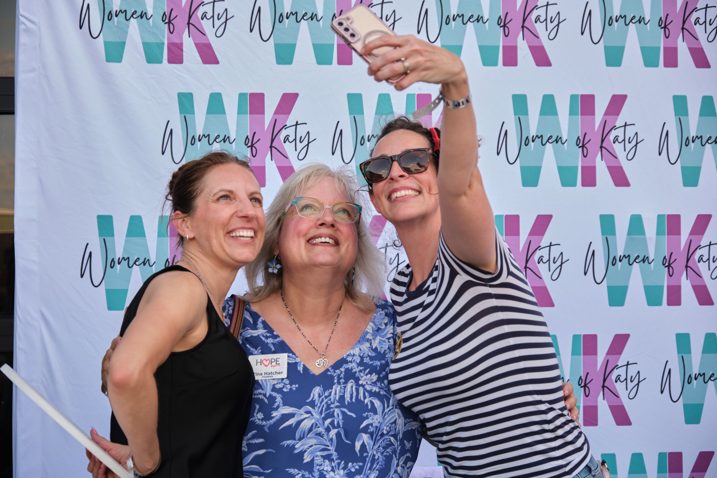 Three women take a selfie together in front of a Women of Katy branded step-and-repeat banner.