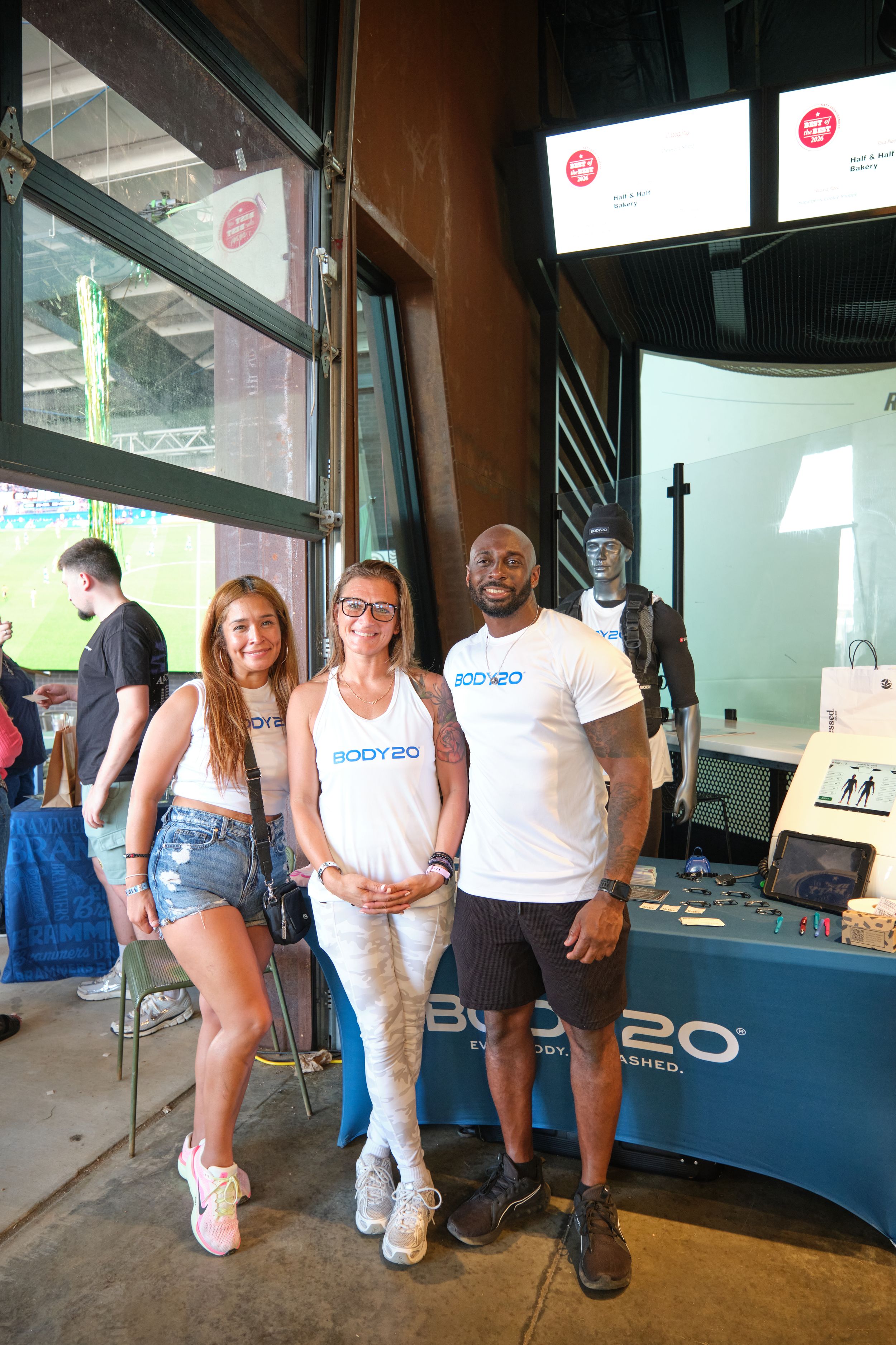 Three people pose together at a Body20 fitness brand booth inside a modern sports venue.