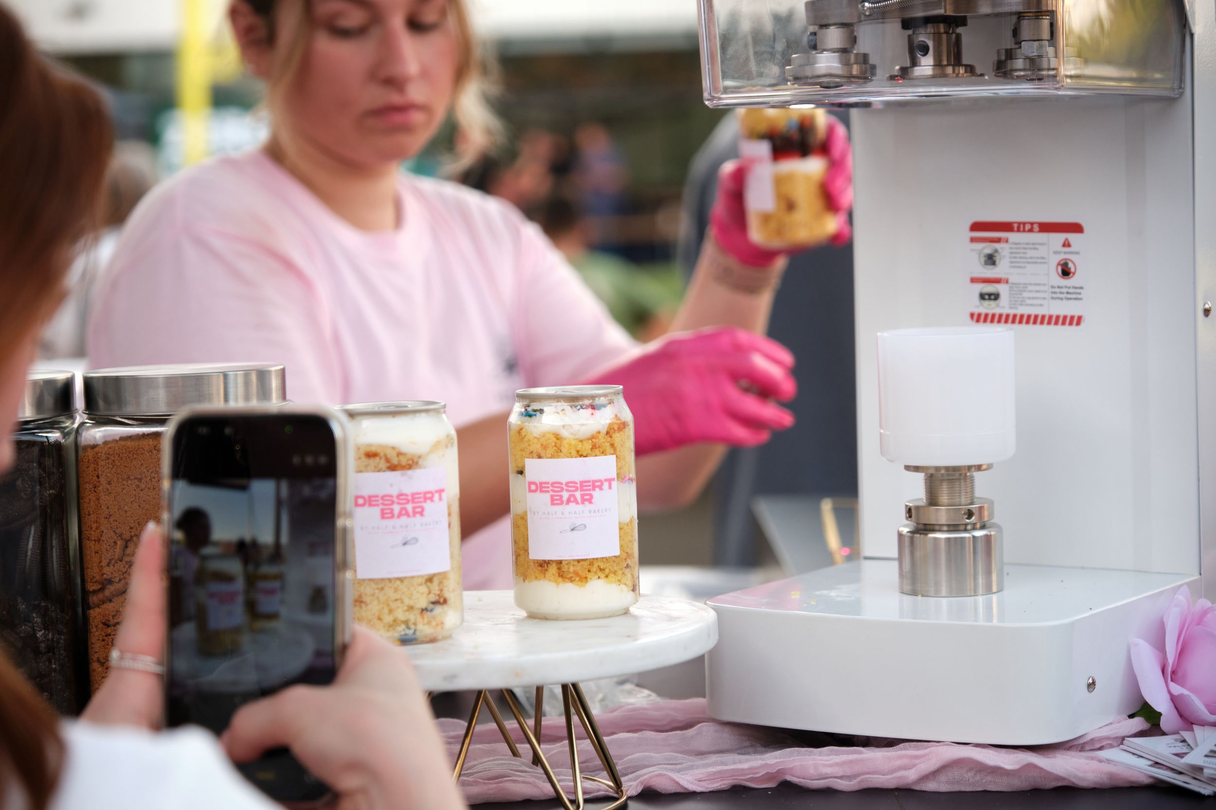 A vendor in pink gloves demonstrates a cookie jar dessert product at an outdoor market booth.